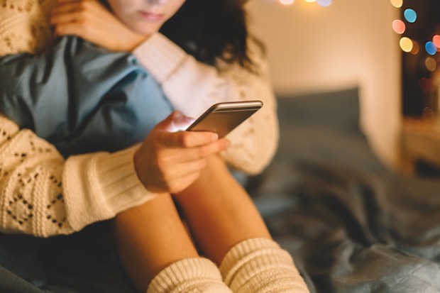 Girl sitting on bed with knees up to their chest using phone.