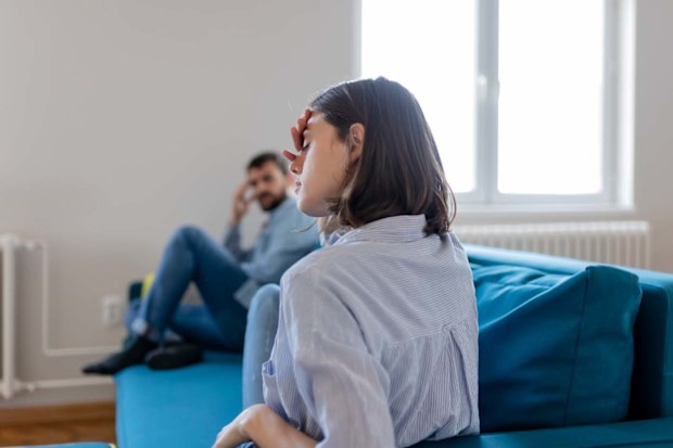 Stressed woman in the foreground sitting on a couch with her hand in her head, he partner sits on the end of the couch looking at her with his hand on his head.
