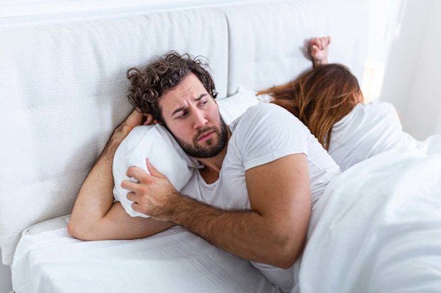 Man and woman lying in bed. The man is in front and looking suspicious of the wife, who is turned the other way, asleep.