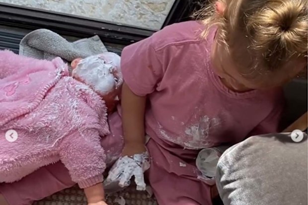 Jen Hawkin's daughter Frankie sitting with her right hand covered in sudocream, holding the container in her left hand, and with her doll on her right with a face covered in sudocream.
