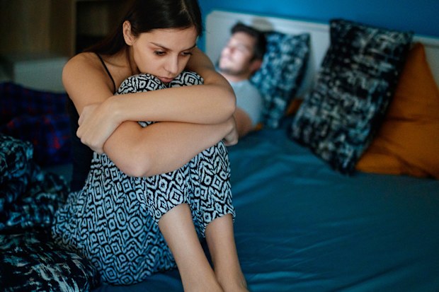 Sad girlfriend sitting on bed edge, holding her legs after fight, her partner is lying in bed in background, his face is lit by smart phone