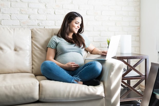 Full length of Caucasian pregnant female using laptop while sitting on sofa in living room
