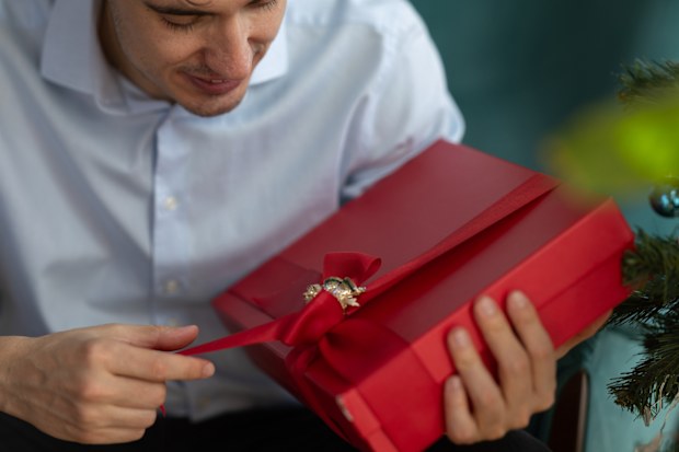 Young man sitting on a vibrant green sofa, joyfully opening a christmas present wrapped with a bright red ribbon, surrounded by a beautifully decorated christmas tree