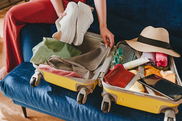 Beautiful smiling young woman packing personal belongings in a suitcase for travelling looking at list at tablet sitting on the sofa in the living room.