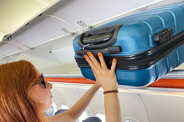 Stock photo showing close-up view of row of carryon luggage overhead baggage compartments in an airplane cabin with beautiful red haired woman putting away her suitcase.
