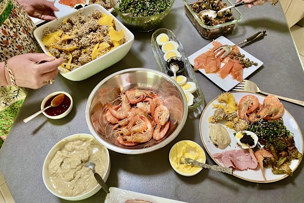 Horizontal spread of a variety of Christmas lunch delicacies on bench in domestic kitchen with hands serving and filling plates including fresh seafood of prawns oysters smoked salmon ham dips tabouli meatballs eggs and more