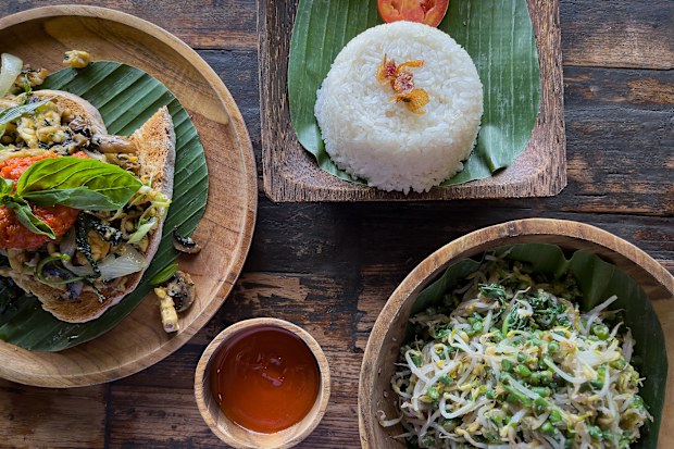 Indonesian-Western fusion breakfast: toast with tempeh, spicy tomato sauce, grilled vegetables, mushrooms, garnished with basil leaves.  Jukut Urab (green beans, spinach and sprouts) served with rice and sambal.  Zero waste leaves, wood plate, bowl, cup and table.  Ubud, Bali, Indonesia.