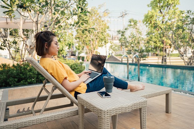 Freelancer working with laptop and smart phone and lying on deck chair near the pool at resort on summer vacation.