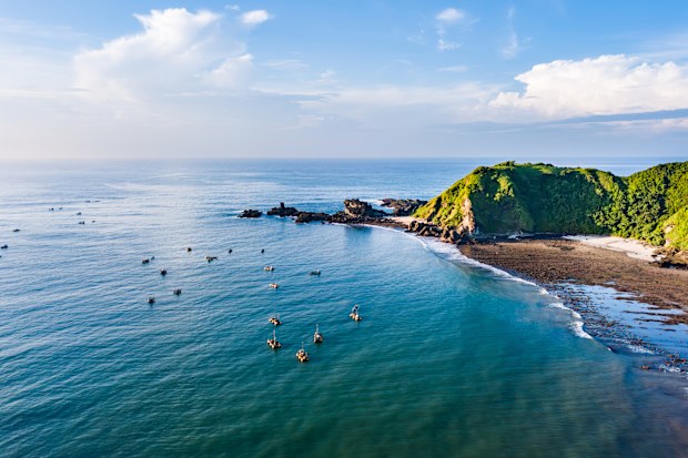 A lush green hillside meets the clear blue waters of the ocean. The rocky shoreline is visible, adorned with various shapes and sizes of rocks. In the distance, a cloudy sky paints a serene backdrop. Shot taken on Lombok.