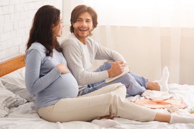 Young pregnant couple choosing name for their future baby, discussing and writing options to notebook, sitting on bed at home together, free space