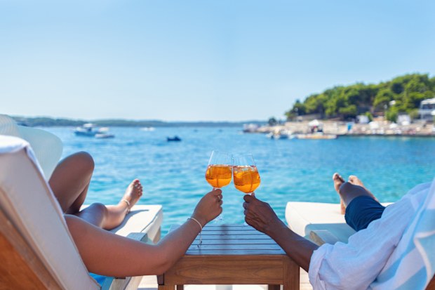 Couple relaxing and toasting with a Aperol spritz cocktail on a beach deck over the ocean. Turquoise ocean is crystal clear.  horizon in the distance.