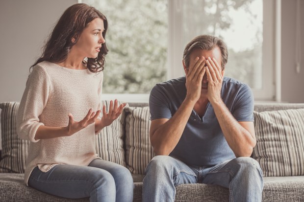 Couple of adults are quarreling while sitting on couch at home, man is covering his face