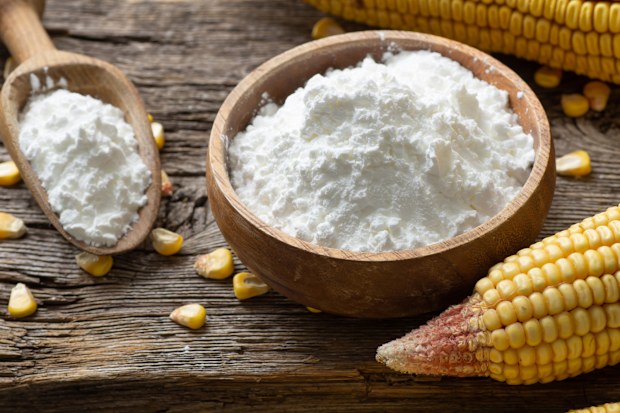 Corn starch, cornflour in bowl and spoon with dried corn groats, kernels on rustic table. Maize starch with ripe cobs and kernels