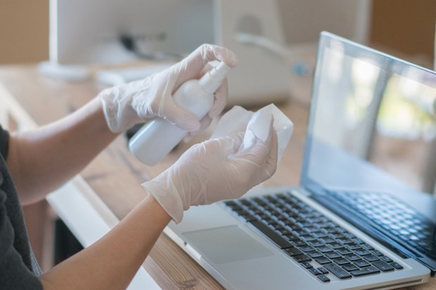 Closeup of the hands of a person disinfecting a laptop's keyboard and screen.