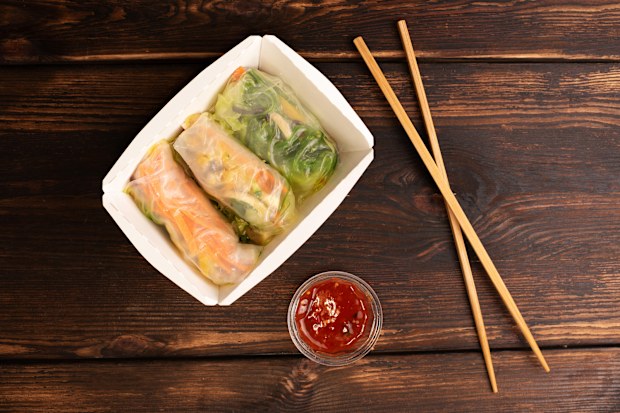 Three spring rolls in paper box with chopsticks and sauce on wooden background, asian fast food, street food concept, close up.