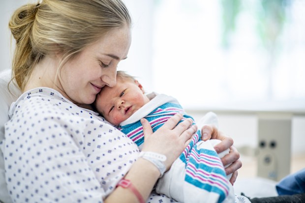 A caucasian mother in a hospital gown sits up in bed and holds her newborn baby to her chest