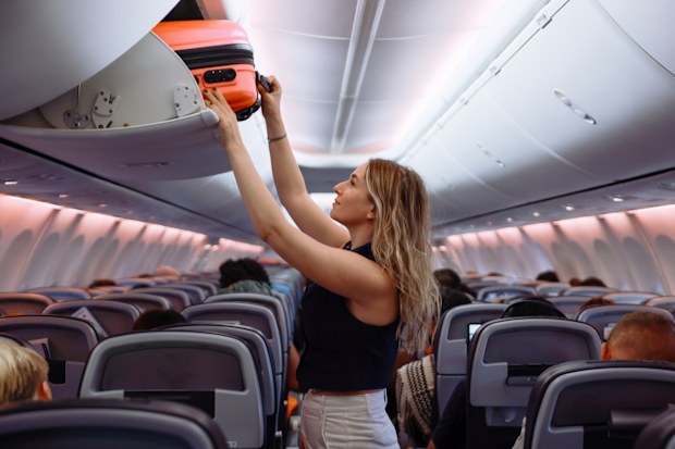 Woman travel by airplane. Passenger putting hand baggage in lockers above seats of plane.  hands are seen placing or retrieving a pink hard-shell suitcase from an airplane's overhead storage compartment.