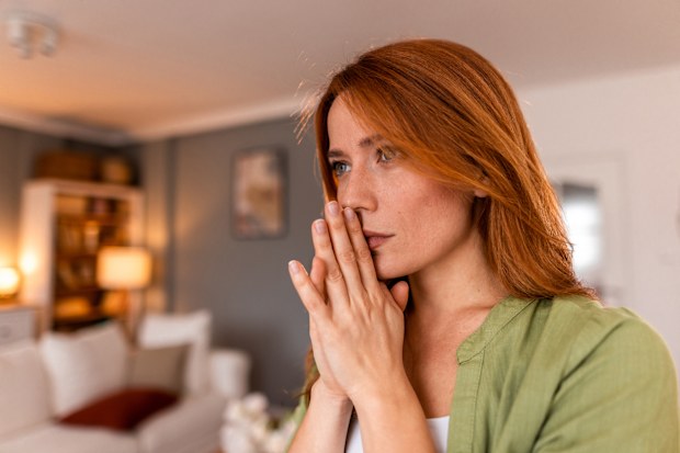 Thoughtful redhead woman with hands clasped, gazing away while standing in a softly blurred living room, reflecting on feelings of solitude and uncertainty in a peaceful home setting