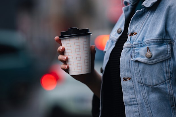 Close up of an unrecognizable woman in denim jacket, walking outside and holding a paper coffee cup to go.