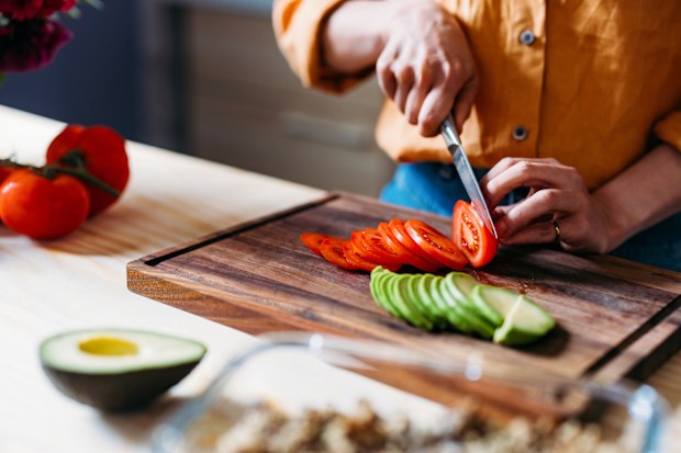 Close-up of hands slicing a tomato and avocado on a wooden cutting board in a kitchen.