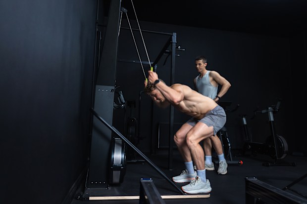 A wide shot of two young men exercising with a SkiErg machine in a modern gym. One man is wearing grey shorts and sports shoes; he performs a resistance training movement targeting strength and muscle definition. The other man stands to the side and watches. Videos are available similar to this scenario.