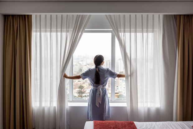 Rear view of a maid working at a hotel and opening the drapes in a room while looking through the window
