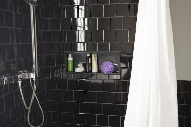 A shot of a black-tiled bathroom with bathroom toiletries on a marble shelf and a shower faucet and a curtain.
