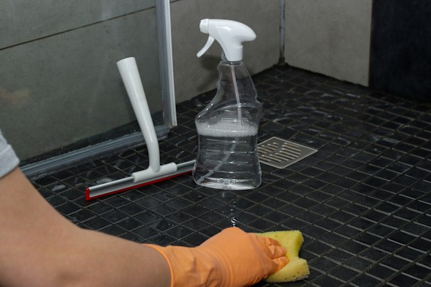 Close up of woman's hand in orange gloves cleaning dirty shower floor tiles with lime remover