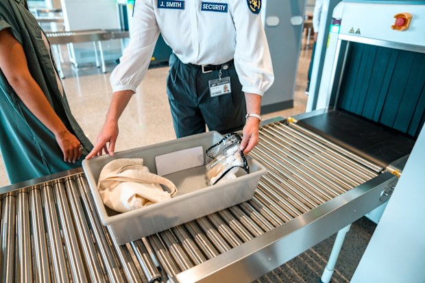 Female security employee holding a plastic tray with  female traveler's personal items, before pushing it through the baggage scanner. Only arms of women visible.