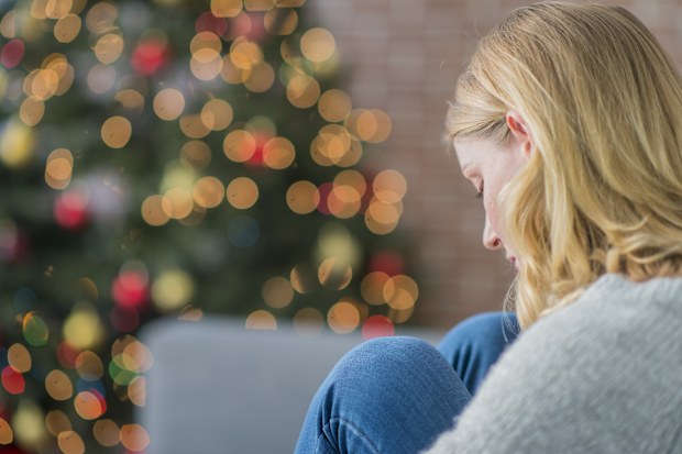 A Caucasian woman is indoors in her living room. There is a Christmas tree in the background. The woman is wearing warm clothing. She is sitting on the couch and looking sad because she is alone on Christmas day.