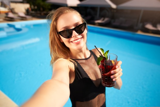 Travel blogger woman in bikini taking selfie photo with cocktail near swimming pool. Pov - lifestyle vlogger selfie from vacation on luxury resort. Female in swimwear drinks beverage on tropical spa.