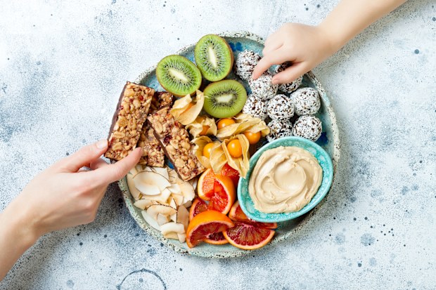 Mother sharing healthy vegan dessert snacks with toddler child. Concept of healthy sweets for children. Protein granola bars, homemade raw energy balls, cashew butter, toasted coconut chips, fruits platter