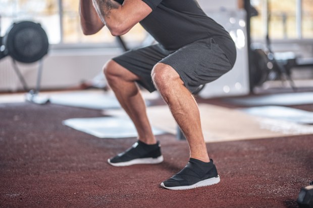 Full length of confident male athlete doing squats at gym. Mid adult man is training at health club. He is in sports clothing.