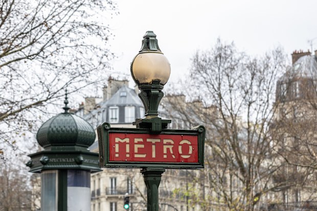 A traditional green Metro sign in the Val d'Osne style, with a vintage Morris column for advertising visible in the background. A concept of historic Parisian street furniture and public life.