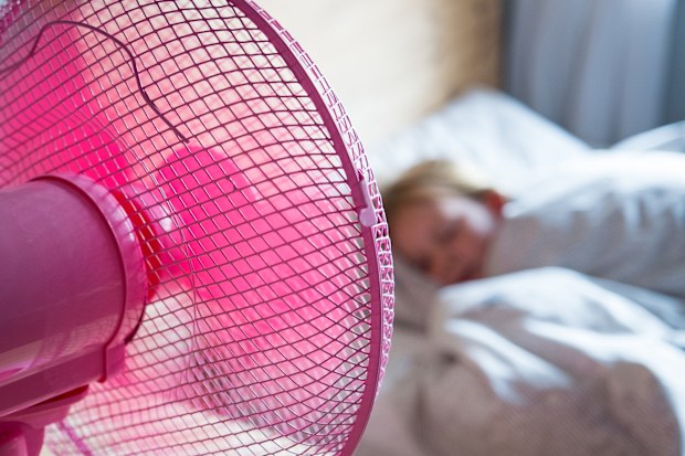 A young girl sleeps in her bed and has a pink fan on her desk blowing cool air over her.