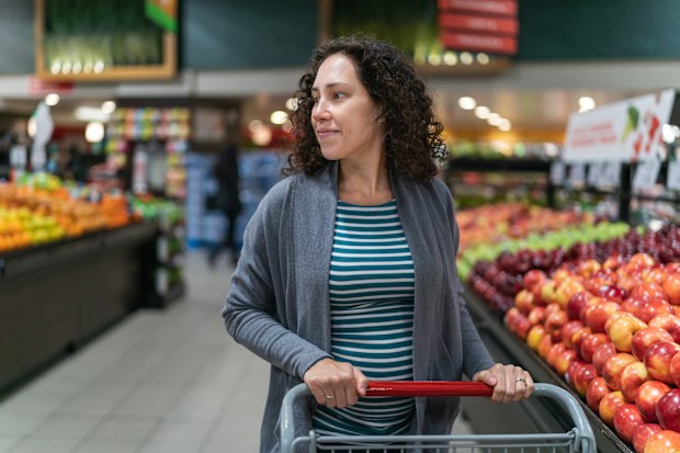 A young mixed race pregnant woman is at the grocery store. She is buying fresh fruit and vegetables in the produce section of the market. The healthy woman is smiling while pushing her grocery cart next to the apples. She is wearing casual maternity clothing.