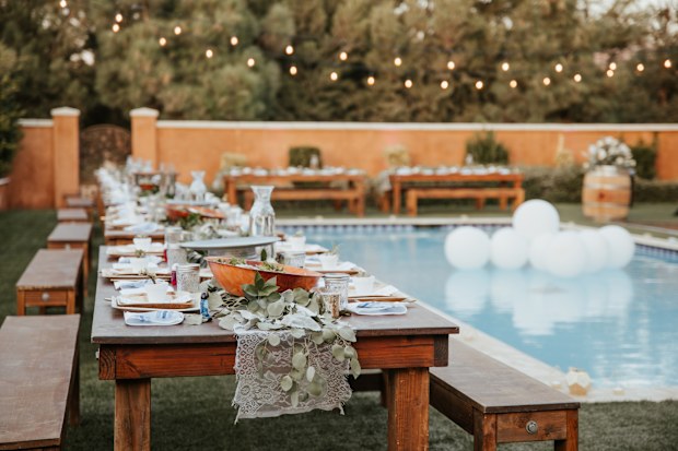Backyard wedding setting - wooden tables with bohemian decor around swimming pool.