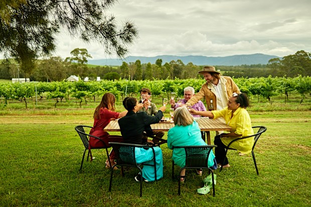 Vineyard host leaning in to touch glasses with table of men and women enjoying rosé selection outdoors with estate and Broken Back Range in background.