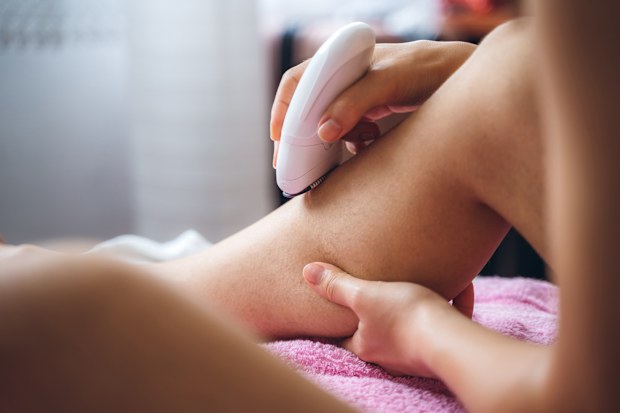 Young caucasian slim woman shaving legs with electric razor in bedroom.