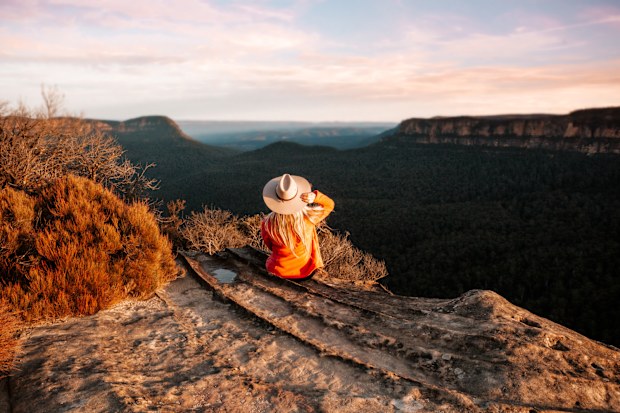 Woman sits on the edge of a cliff and looks out over the mountains in teh late afternoon