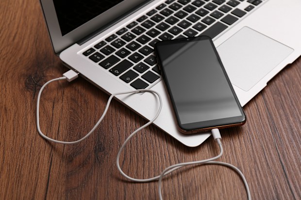 Smartphone connected with charge cable to laptop on wooden table. Space for text