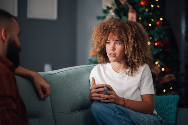 Worried young woman is holding a mug and having a serious conversation with her husband while sitting on a sofa during christmas holidays