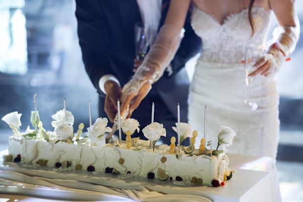 Newlyweds happily cut, laugh and taste the wedding cake. Beautiful Bride and Groom Celebrate Wedding at an Evening Reception Party with Multiethnic Friends. Married Couple Standing at a Dinner Table, Cutting Wedding Cake.