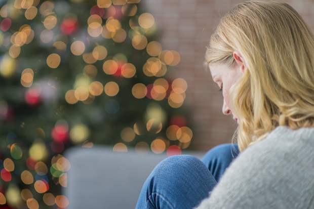 A Caucasian woman is indoors in her living room. There is a Christmas tree in the background. The woman is wearing warm clothing. She is sitting on the couch and looking sad because she is alone on Christmas day.