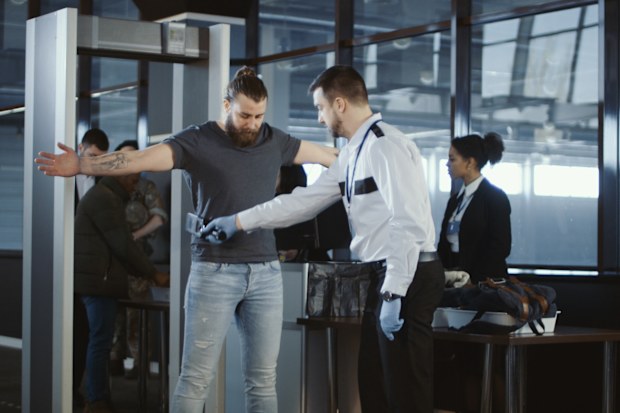 Security agent at an airport check-in gate patting down a bearded casual male passenger with outstretched arms after he passes through the metal detector scanner in the departures hall.