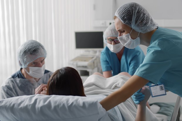 Woman giving birth with husband holds her hand in support and obstetricians assisting. Back view of medical staff in protective uniform helping pregnant woman in labor