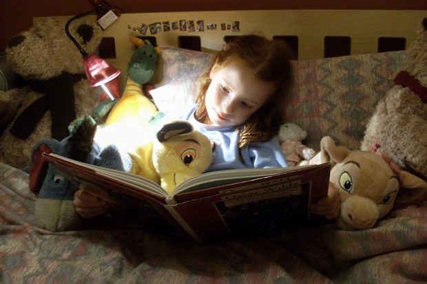 Frome, Somerset, England, UK - March, 16 2009: Candid photo showing a young girl reading a book at home, in bed by the light of a reading lamp, surrounded by cuddly toys.