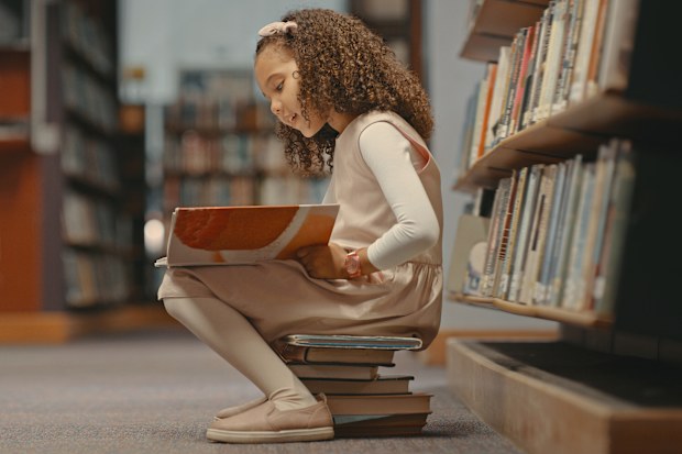Young girl sitting on books in the library and reading a book. Cute girl with curly hair doing her project. Female alone and doing research for a project
