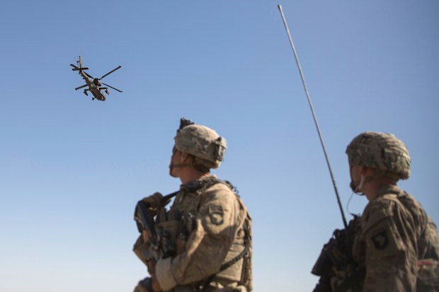 An AH-64 Apache attack helicopter flies above as US Soldiers at Task Force Iron at Bost Airfield in Afghanistan watch on.