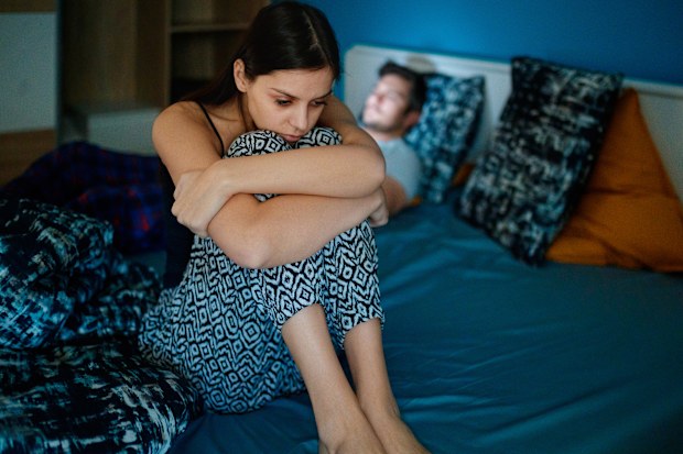 Stock image of a woman sitting on a bed's edge while her partner is lying in bed in the background.